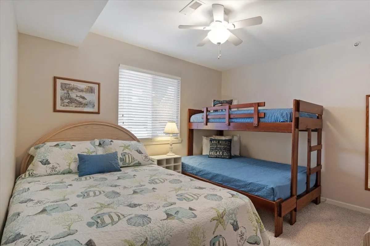 A family-friendly bedroom with both a double bed featuring a playful fish-patterned quilt and a bunk bed with blue bedding. The room includes a table lamp and framed artwork, creating a cozy and inviting vibe for children or families.