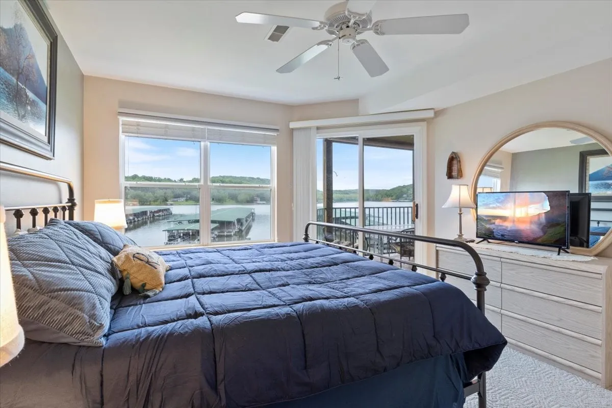 A peaceful bedroom with a bed featuring a soft blue comforter, large windows that let in natural light and reveal calming lake views, a round mirror above a dresser, and a television for relaxing evenings in bed.