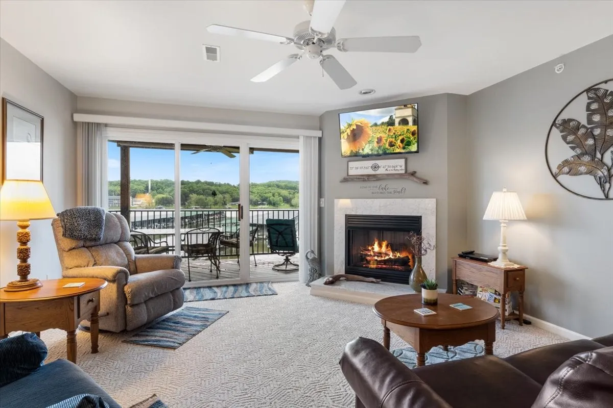 A warm living space featuring a fireplace, large windows, a TV, and a spacious seating area. The arrangement invites relaxation, reading, or group entertainment, with ample natural light and a glimpse onto an outdoor balcony.
