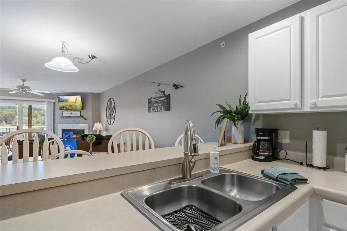A welcoming interior focalizing on the kitchen sink and dining table, with expansive windows framing a picturesque lake view. The decorative wall sign adds a touch of homey charm. The space is set up for cooking, entertaining, and enjoying meals with family or friends while taking in the scenery.