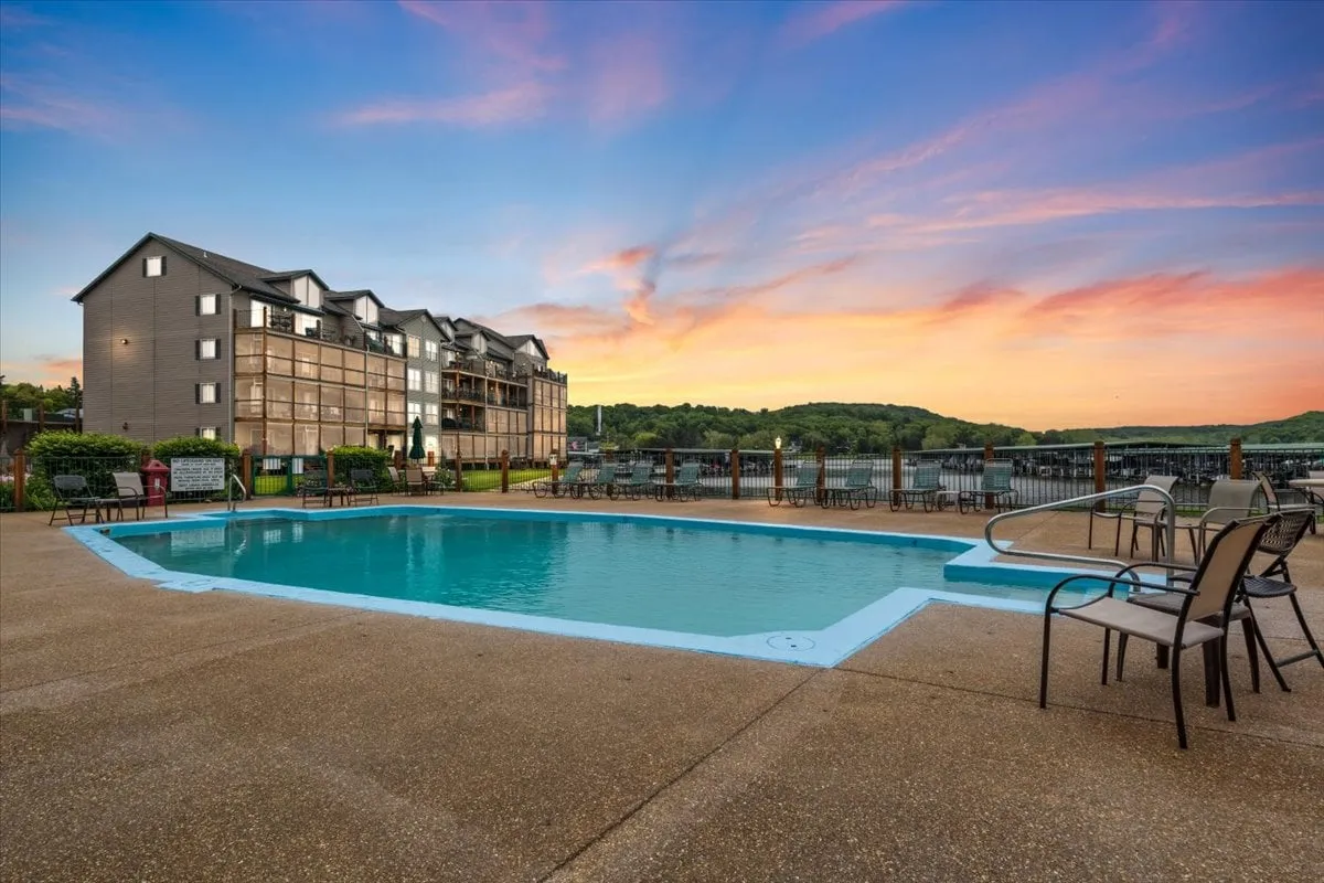 The image showcases a sparkling swimming pool at sunset, with inviting lounge chairs and a view of the calm lake beyond. The surrounding condo buildings are visible, creating a friendly, resort-like atmosphere, while the tranquil sky casts a warm, peaceful glow over the entire scene. The pool area is perfect for both relaxing swims and social gatherings, suggesting a mix of family fun and serene getaways.