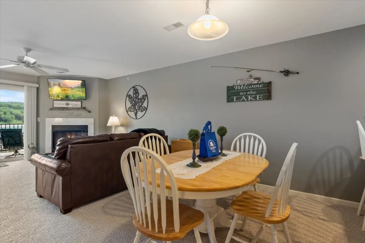 A cozy dining area with a welcoming table is set beside a fireplace, and through large windows, tranquil lake views are visible. The 'welcome' sign and comfortable seating add a sense of arrival and relaxation. Sunlight streaming in hints at the condo's direct visual connection to the water, providing a sense of lakeside ambiance from indoors.