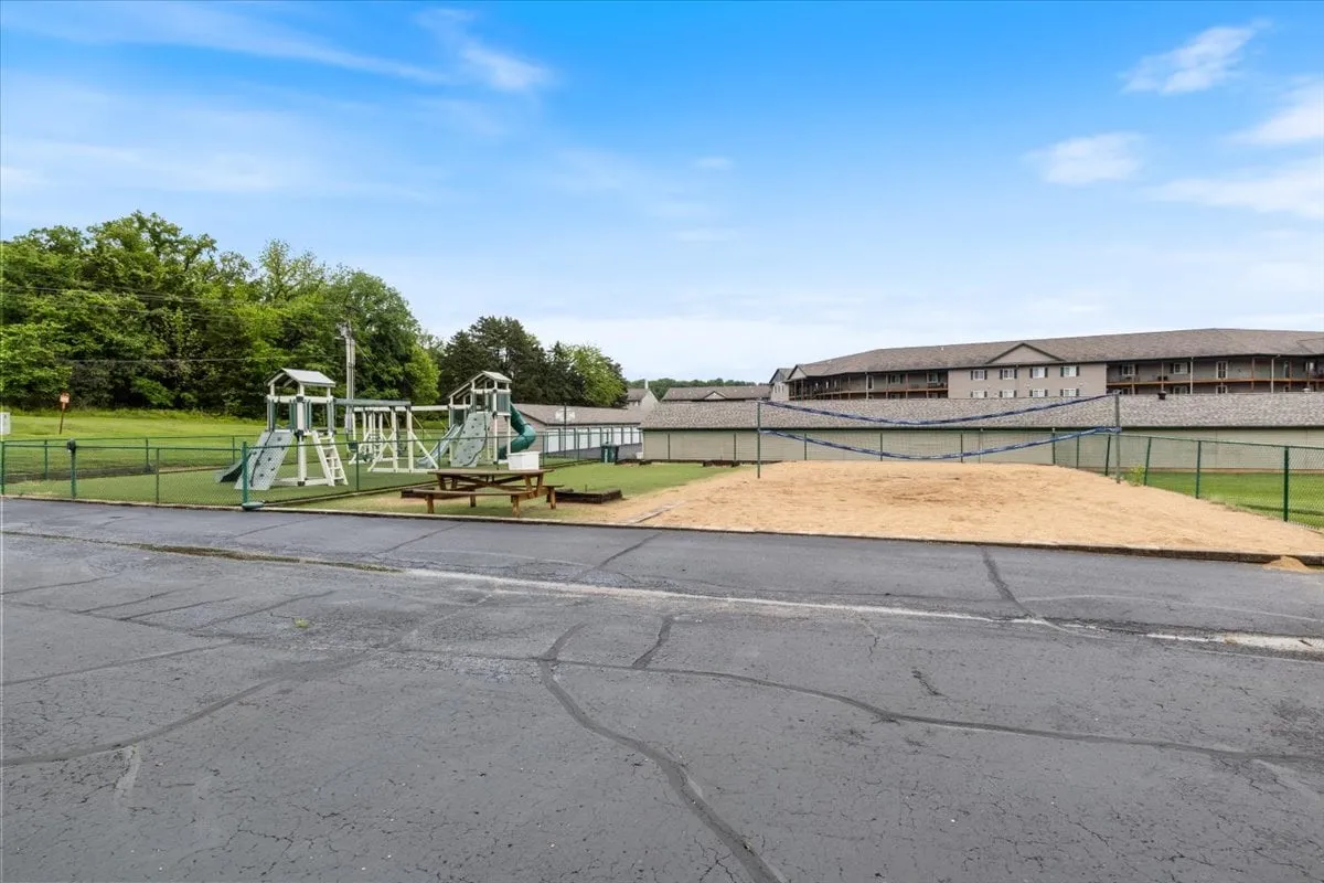 A lively outdoor playground with swings and climbing structures, a sand volleyball court, and a lush green lawn where children play and families gather. The image radiates a joyful and active community vibe.