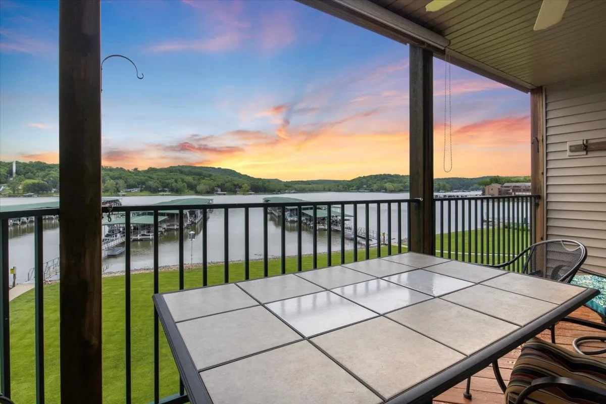 A scenic view from the private deck, highlighting an outdoor dining table positioned to enjoy expansive lake views. The sun is setting, bathing the sky and landscape in warm hues, perfect for both relaxing mornings and celebratory evenings outdoors.